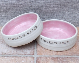 May include: Two white ceramic pet bowls with pink interiors. The bowls are personalized with the text "Ginger's Water" and "Ginger's Food".