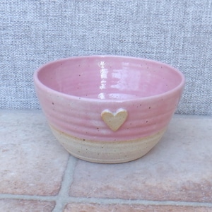 Puede incluir: Un cuenco de cerámica hecho a mano con un esmalte rosa y una base natural sin esmaltar. El cuenco presenta un pequeño detalle en forma de corazón. El interior es rosa brillante y el exterior tiene un acabado mate texturizado. Aproximadamente 15 cm de diámetro.