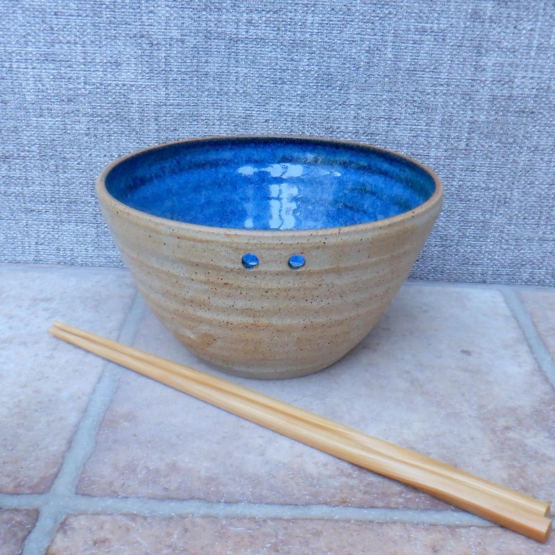 May include: A handcrafted ceramic bowl with a bright blue interior and a tan exterior. Two small holes are visible on the side. A pair of wooden chopsticks rests beside the bowl, suggesting its use for Asian cuisine.