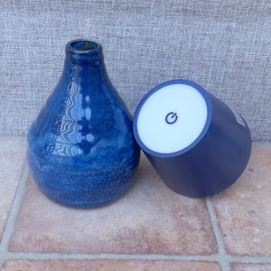 May include: A glossy, deep blue ceramic vase with a textured, rippled surface next to a dark blue cylindrical object featuring a white top panel with a black power button symbol printed in the centre. Both items are positioned side by side.