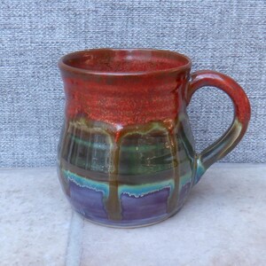 May include: A handcrafted ceramic mug with a layered glaze in red, green, blue, and purple. The mug features a rounded body and a curved handle. The glaze has a dripped effect, adding to its unique character.