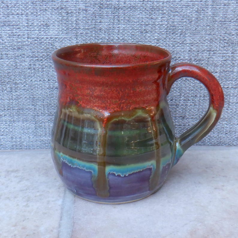 May include: A handcrafted ceramic mug with a layered glaze in red, green, blue, and purple. The mug features a rounded body and a curved handle. The glaze has a dripped effect, adding to its unique character.