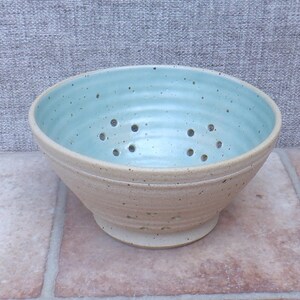 May include: A ceramic berry bowl with a light blue interior and a speckled beige exterior. The bowl features a series of drainage holes arranged in a circular pattern along the sides and base, with a ribbed texture on the outer surface.