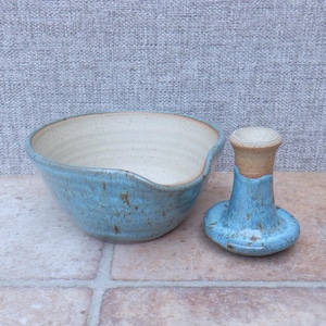 May include: A ceramic mixing bowl with a light blue speckled exterior and a cream-colored interior, featuring a small pouring spout on the rim. Beside the bowl sits a matching ceramic pestle with a blue speckled base and a cream-colored handle.