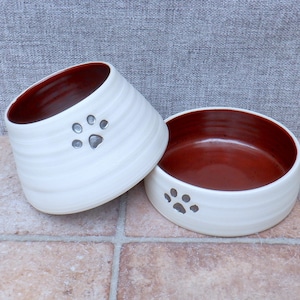 Puede incluir: Dos cuencos de cerámica blanca para mascotas con interior rojo y diseño de huella de pata plateada.