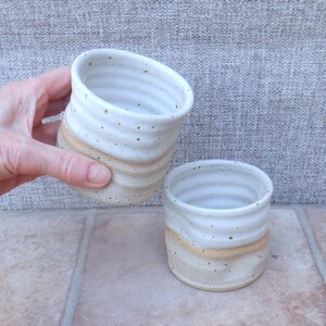 Puede incluir: Dos tazas de cerámica hechas a mano. Las tazas son blancas con una banda beige en el medio y salpicadas con puntos marrones. Una taza es sostenida por una mano. Las tazas tienen una textura acanalada.