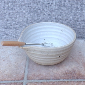 Puede incluir: Un cuenco de cerámica beige con un pico vertedor y un pequeño batidor con mango de madera. El cuenco tiene un exterior texturizado y un interior moteado. El batidor es de acero inoxidable.