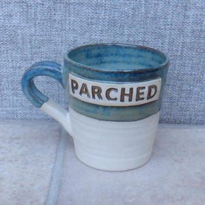 May include: A handmade ceramic mug with a blue rim and handle, and a white base. The word "PARCHED" is printed in brown letters on a white rectangular label on the mug's side. The mug is on a light grey surface.