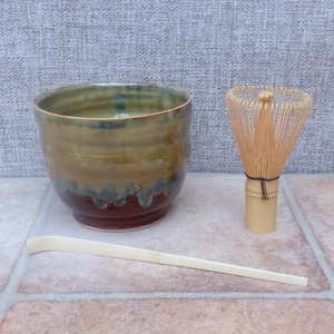 May include: A ceramic matcha bowl with a brown, green, and blue glaze, a bamboo whisk, and a bamboo scoop. The bowl has a wide mouth and a rounded shape. The whisk is made of thin bamboo tines. The scoop is long and flat.