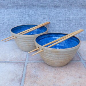 May include: Two handmade ceramic bowls with a textured, tan exterior and a vibrant blue interior. Each bowl holds a pair of wooden chopsticks. The bowls are designed with a groove to hold the chopsticks. The bowls are placed on a tiled surface.