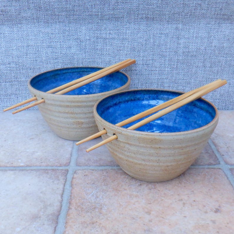 May include: Two handmade ceramic bowls with a textured, tan exterior and a vibrant blue interior. Each bowl holds a pair of wooden chopsticks. The bowls are designed with a groove to hold the chopsticks. The bowls are placed on a tiled surface.