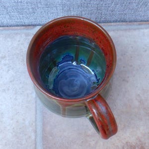 May include: Handmade ceramic mug with a unique glaze. The interior features a deep blue swirl, transitioning to green and then a vibrant red rim. The handle and exterior have a reddish-brown hue, creating a rustic, artistic look.