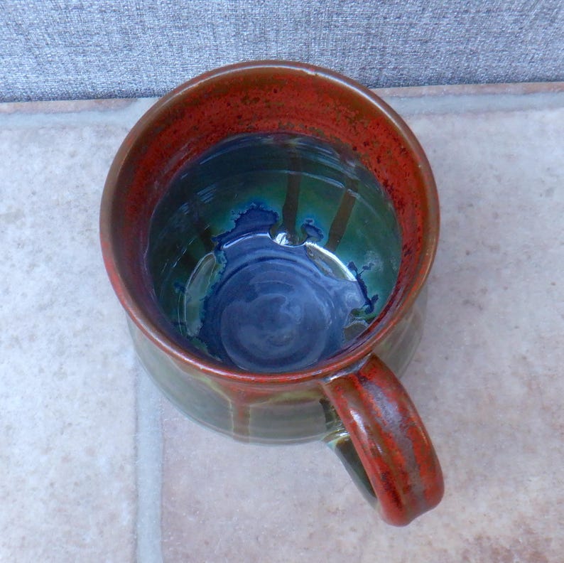 May include: Handmade ceramic mug with a unique glaze. The interior features a deep blue swirl, transitioning to green and then a vibrant red rim. The handle and exterior have a reddish-brown hue, creating a rustic, artistic look.