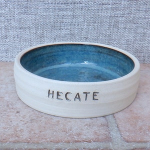 Puede incluir: Tazón de cerámica blanco para mascotas con un esmalte azul en el interior. El tazón tiene el nombre "HEcate" inscrito en la parte delantera.