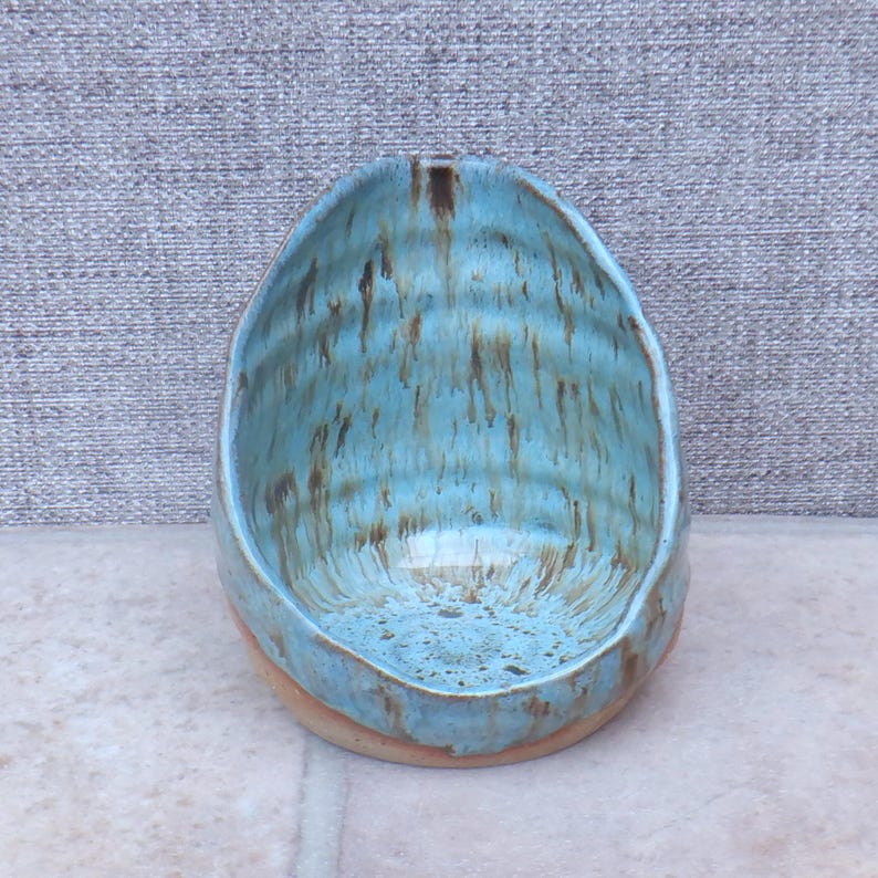 May include: A handcrafted ceramic bowl with an irregular form. It has a light blue glaze with brown streaks, giving a textured, artistic look. The base is a natural, unglazed terracotta colour.