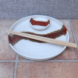 May include: A white ceramic sushi plate set with a small dipping bowl and chopsticks. The plate and bowl have a brown and red painted design. The chopsticks are light brown.