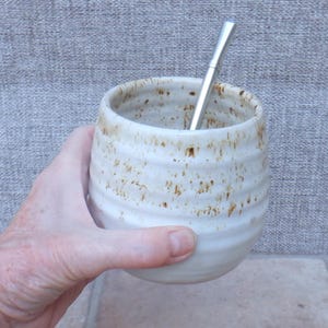 May include: A round, off-white ceramic cup with horizontal ridges and brown speckled detailing. The cup features a wide opening and a rounded base, accompanied by a slender, straight silver-colored metal straw inserted into the top opening.
