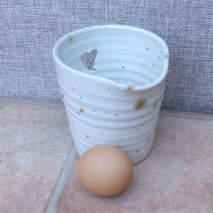 Puede incluir: Un huevo de cerámica blanco con un recorte en forma de corazón y un glaseado moteado marrón y azul. Un huevo marrón se encuentra delante de la taza.