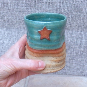 Könnte beinhalten: Handgefertigte Keramiktasse mit türkisfarbenem Oberteil, braunem Stern und orangefarbenem Band. Der Boden ist beige mit einem strukturierten, geriffelten Design. Ein einzigartiges, handgefertigtes Stück für Getränke.