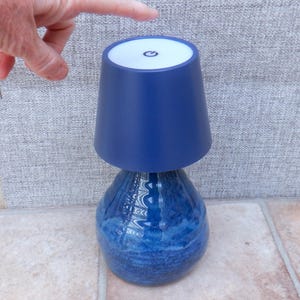 May include: A blue table lamp with a textured, bulbous base and a smooth, tapered shade. The top of the shade features a white circular panel with a black power symbol icon centred on it, indicating a touch-sensitive control mechanism.