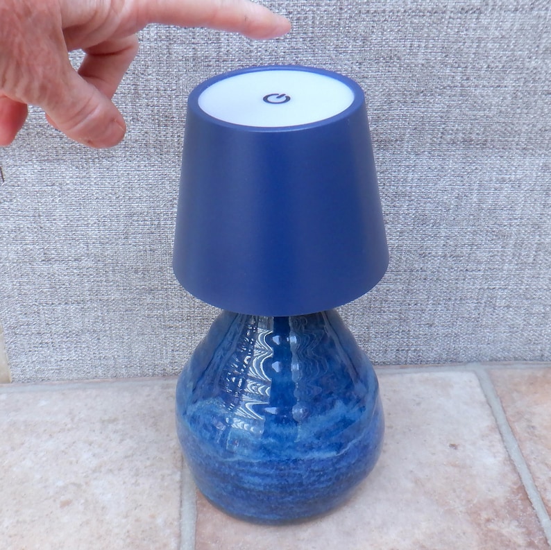 May include: A blue table lamp with a textured, bulbous base and a smooth, tapered shade. The top of the shade features a white circular panel with a black power symbol icon centred on it, indicating a touch-sensitive control mechanism.