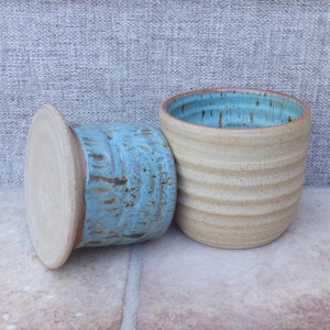 May include: Two handmade ceramic containers. One is a light beige colour with horizontal ridges and a turquoise interior. The other is a matching container with a turquoise exterior and a light beige lid.