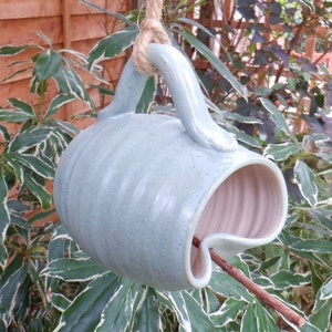 May include: A light blue ceramic bird feeder shaped like a mug with a rope hanging loop. The feeder has a small opening for birds to access the food.