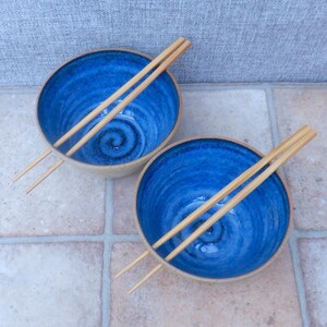 May include: Two ceramic bowls with a striking blue interior and a natural beige rim. Each bowl has a spiral design and comes with wooden chopsticks. These bowls are suitable for serving soup or noodles, a stylish addition to any kitchen.