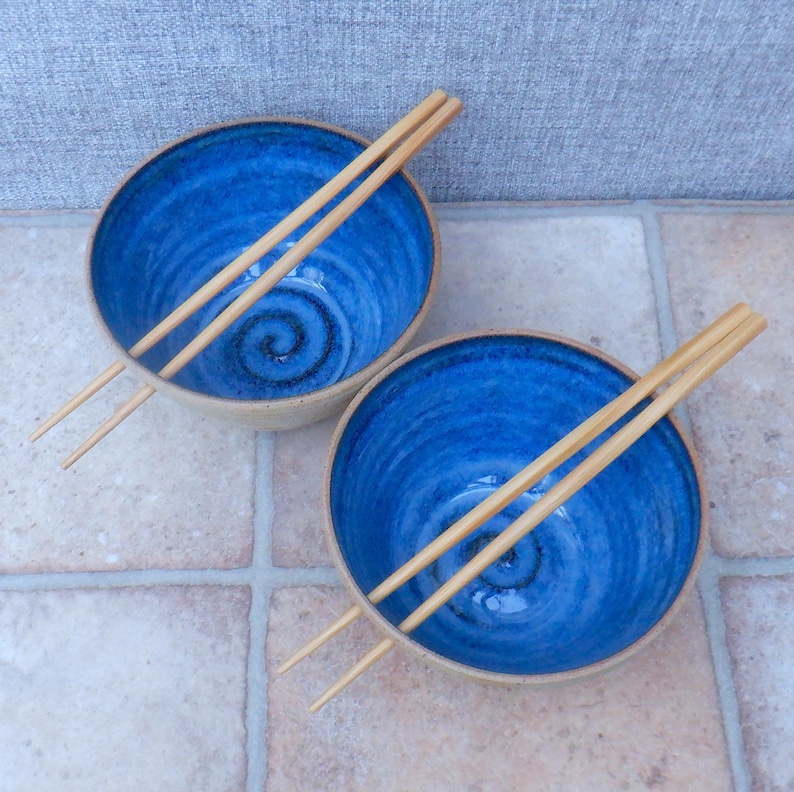 May include: Two ceramic bowls with a striking blue interior and a natural beige rim. Each bowl has a spiral design and comes with wooden chopsticks. These bowls are suitable for serving soup or noodles, a stylish addition to any kitchen.