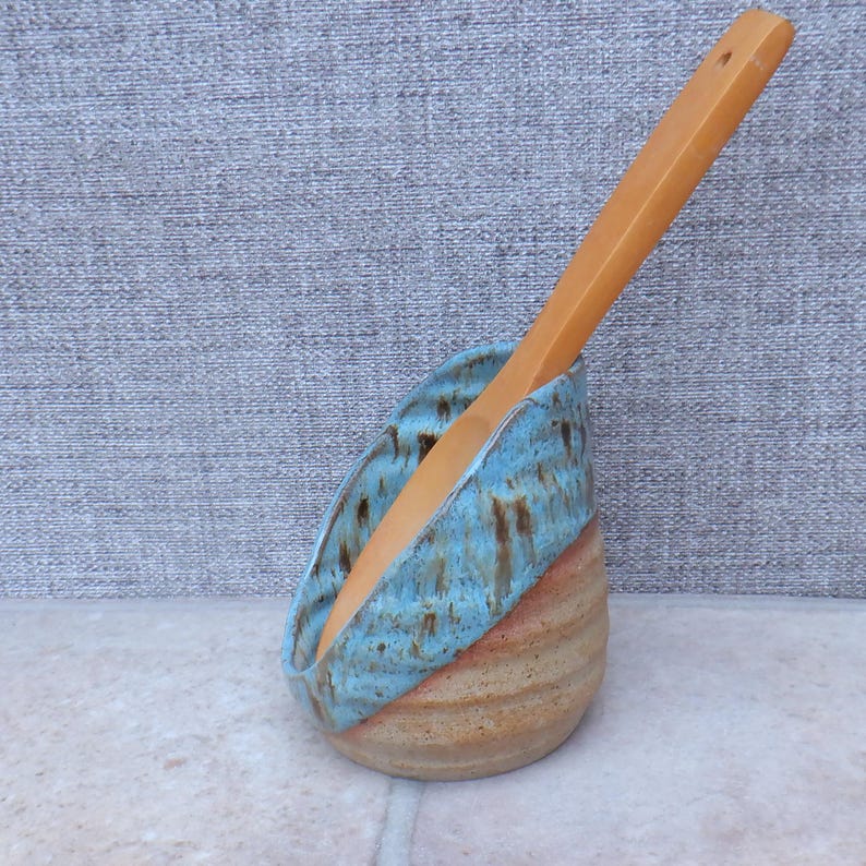 May include: A ceramic utensil holder with a wooden spoon. The holder has a light blue glazed rim over a tan and brown striped body. The wooden spoon is light brown and is placed inside the holder. The holder is handmade.
