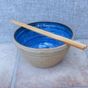 May include: A handcrafted ceramic bowl with a striking blue interior and a light brown exterior. The bowl is accompanied by wooden chopsticks, ideal for serving Asian cuisine. The bowl has a diameter of approximately 20 cm.