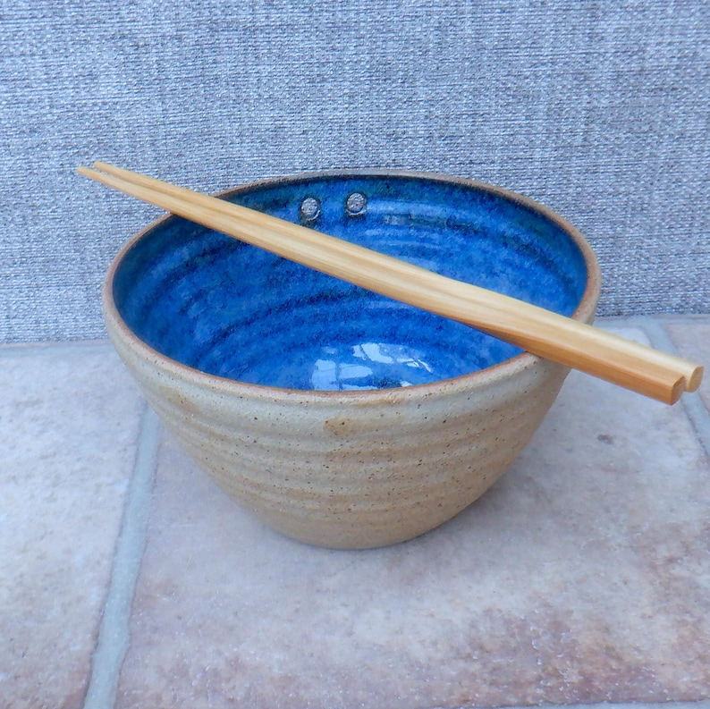 May include: A handcrafted ceramic bowl with a striking blue interior and a light brown exterior. The bowl is accompanied by wooden chopsticks, ideal for serving Asian cuisine. The bowl has a diameter of approximately 20 cm.