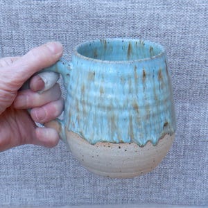 Può includere: Una tazza in ceramica fatta a mano con una forma arrotondata e un grande manico. La tazza presenta una glassa azzurra con striature marroni e una base naturale non smaltata. La tazza è tenuta da una mano.