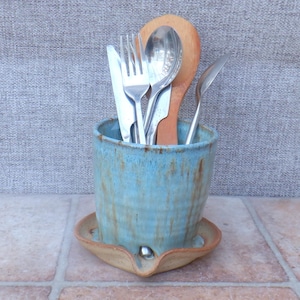 May include: A handmade ceramic utensil holder in a light blue glaze with brown accents. The holder contains silverware and a wooden spoon. The holder sits on a matching saucer with a small spout.