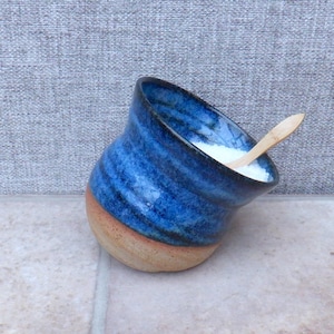 Puede incluir: Un salero de cerámica hecho a mano con exterior esmaltado azul y base beige natural. El interior contiene sal blanca y una pequeña cuchara de madera. El diseño inclinado añade un toque único a este utensilio de cocina.