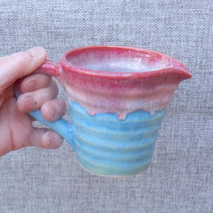 May include: A handmade ceramic pitcher with a blue and pink glaze. The pitcher has a wide mouth and a handle. The glaze is uneven, creating a unique and rustic look.
