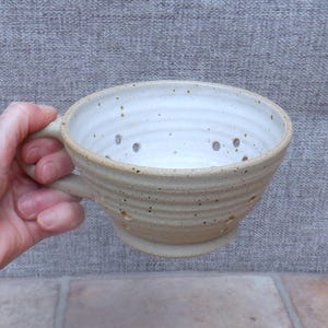 May include: A handmade ceramic colander with a handle. The colander is beige with a white interior and small drainage holes. The colander is designed for straining liquids from food items.