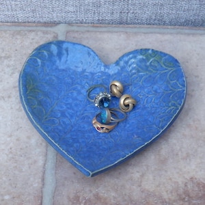 Puede incluir: Un plato de cerámica azul en forma de corazón con un patrón de hojas. El plato está lleno de cuatro anillos, incluyendo dos anillos de oro y dos anillos con piedras preciosas azules.