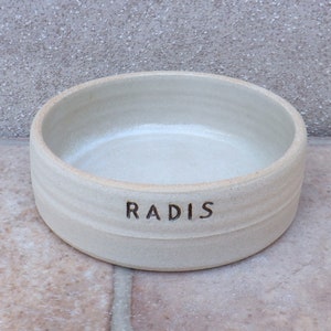 Puede incluir: Un cuenco de cerámica blanco para mascotas con la palabra "RADIS" inscrita en la parte delantera.