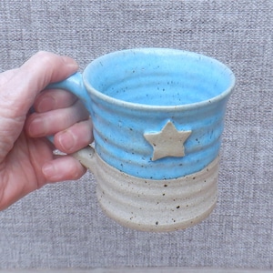 Puede incluir: Una taza de cerámica hecha a mano con una parte superior esmaltada azul claro y una parte inferior sin esmaltar de color beige. Una estrella beige está adherida a la sección azul. La taza está sujeta por una mano.