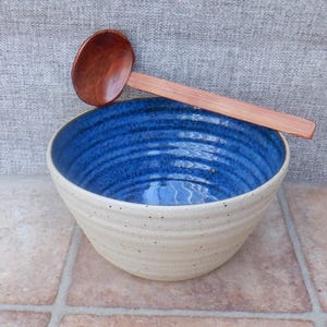 Puede incluir: Un cuenco de cerámica redondo con exterior crema moteado e interior azul profundo, con estrías horizontales en los lados. Un cucharón de madera de mango largo con un acabado liso y pulido descansa sobre el borde superior del cuenco.