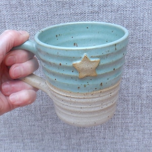 Könnte beinhalten: Handgefertigte Keramiktasse mit hellblauem Oberteil und beigem Unterteil. Die Tasse hat eine sternförmige Verzierung und eine gerippte Textur. Der Griff und der Stern sind beige. Die Tasse wird von einer Hand gehalten.