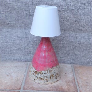 May include: A table lamp featuring a conical ceramic base with a vibrant pink glaze on the upper portion and a speckled beige finish on the lower section. The lamp is topped with a simple, solid white conical shade that tapers toward the top.