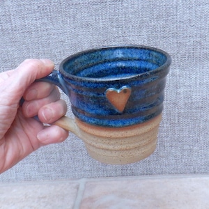 Puede incluir: Taza de cerámica hecha a mano en azul y marrón. La parte superior es azul con rayas azul oscuro, la inferior es marrón con estrías horizontales. Un pequeño corazón marrón está adherido al lateral. La taza tiene un asa curva.