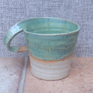 May include: A ceramic mug featuring a two-tone design with a glossy, speckled teal glaze on the upper portion and a textured, matte beige finish on the lower base. The mug has a wide, curved handle that attaches near the rim and the side wall.