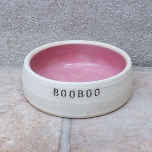 Puede incluir: Tazón de cerámica blanco para mascotas con interior rosa y el nombre "BOOBOO" inscrito en la parte delantera.
