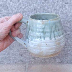 Puede incluir: Una taza de cerámica hecha a mano con un cuerpo redondeado y un asa robusta. La taza presenta un esmalte de dos tonos, con un color azul verdoso claro que cae sobre una base blanca cremosa. La taza es sostenida por una mano.