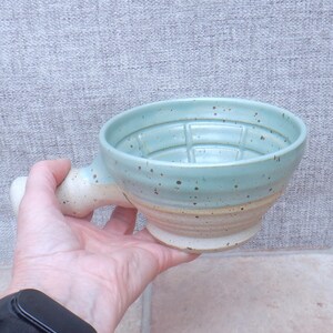 May include: A ceramic shaving bowl with a handle, featuring a light blue glazed interior and upper exterior with brown speckles, transitioning to a speckled beige unglazed base. The interior bottom has raised ridges for lathering soap.