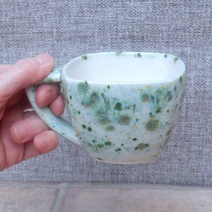 May include: A handmade ceramic mug with a square shape and a handle. The mug is light blue with green and yellow speckled patterns. The interior is white. The mug is held by a hand.