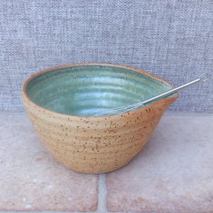 May include: A ceramic mixing bowl with a speckled tan exterior and a smooth, light green interior glaze. The bowl features a ribbed texture along the sides and a small pouring spout. A silver metal wire whisk rests inside the bowl.